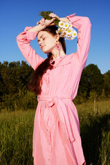 Wall Mural - Stylish young woman in a pink dress with flowers, joyful expression, standing in a green field under a clear blue sky, capturing the essence of summer