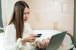 © Ekkasit A Siam - Asian woman in white blouse holding a credit card while looking at laptop with concerned expression. Online transaction, banking, and financial decision-making concept in a modern bedroom setting.