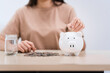 © Phushutter - A young Asian woman in a light brown t-shirt, sitting with a piggy bank, coin, calculator, mobile, and pen. She focuses on financial stability, saving for emergencies, education, and future goals.