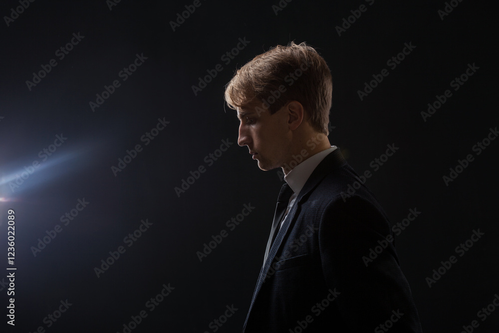 In a dark studio, a man in a black suit and white shirt stands in ...