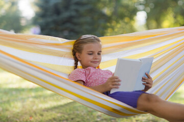 Happy  adorable smiling child girl  lying in a hammock and reads a book in summer park. Happy ghild having fun outdoors. Generation Alpha. copy space. 