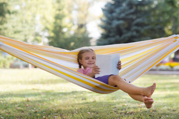 Happy  adorable smiling child girl  lying in a hammock and reads a book in summer park. Happy ghild having fun outdoors. Generation Alpha. copy space. 