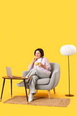 Wall Mural - Mature woman with tea cup sitting in armchair and laptop on table against yellow background