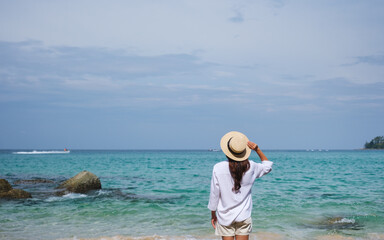 Wall Mural - Rear view image of a woman with hat standing on the beach with blue sky background