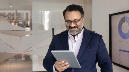 Wall Mural - Happy successful senior Indian business owner man analyzing financial information, marketing report, sales growth, using tablet computer at large electronic board with statistic data. Banner shot