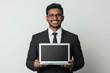 © DLV - Indian man holding laptop. Entrepreneur startup. Remote freelance. Cheerful Indian businessman showing laptop in a professional setting.