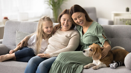 Wall Mural - Beautiful young woman spend priceless time with little daughter and older mother, sit on sofa with dog, exuding family connections, togetherness, intergenerational bonding, relish warm shared moments