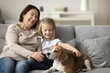 © fizkes - Smiling old woman and little grandchild girl sitting on sofa, gently petting beagle dog, having pleasant time together, exude warmth love, connection between generations, human and pet. Family pastime