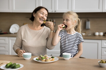 Older grandmother and child eating homemade sandwiches smile look at each other with love and playful expression. Multigenerational family spend time together, enjoy delicious food, and communication