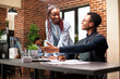 © DC Studio - Smiling black woman brainstorming marketing plans with colleague seated at desk with laptop in startup office. African american employees collaborating and sharing strategy plans in brick wall room.