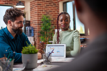 Wall Mural - Black entrepreneur guiding her colleagues with market strategy insights, utilizing a digital device. Female startup leader using a tablet to explain company goals to her team in brick wall office.