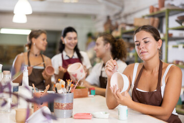 During master class, female participant draws pattern on walls of white ceramic bowl with brush. Girl paints blank, makes individual order. Hand made art, creative pastime.