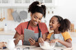 © Prostock-studio - Happy black mummy and daughter cooking at home together, wearing aprons, standing by kitchen table with dough, flour, rolling pin and cookies figurines, using tablet, reading culinary blog