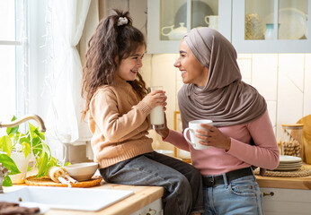 Wall Mural - Happy Muslim Mom In Hijab And Her Little Daughter Bonding Together In Kitchen, Islamic Family Talking And Laughing, Mother Enjoying Coffee While Her Female Child Sitting At Table And Drinking Milk