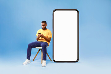 Wall Mural - cheerful young african american man sitting on chair near big smartphone with empty screen, using mo