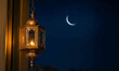 © Masrob - Islamic lanterns hanging near the windows of a beautiful house glow warmly in the dark night sky, with a crescent moon in the dark sky. The image evokes spirituality, Ramadan, and a peaceful nighttime