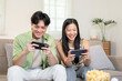 © bongkarn - A cheerful Asian couple enjoys playing a mobile game on their phones, sitting on a sofa together.