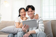 © bongkarn - A happy young Asian couple sits together on a sofa in the living room, watching a video on a tablet.