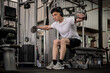© bongkarn - A determined Asian man sits on a fitness bench, working out with dumbbells in the gym.