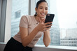 © Malambo20/peopleimages.com - Business, woman and happy with phone by window for checking weather app, work break and online storm update. Professional, employee or smile with smartphone in office for web browsing or reading info