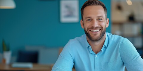 Wall Mural - A man with a blue shirt and a beard is smiling. He is sitting at a table with a laptop and a cup