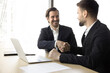© fizkes - Two happy businessmen sit at table with laptop and shaking hands, feel satisfied, express respect, trust, finish meeting in office. Successful conclusion of discussion, business negotiation, agreement