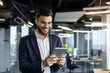 © Liubomir - Successful businessman in business suit holding tablet computer. Man inside office at workplace smiling happily, using financial investment app.