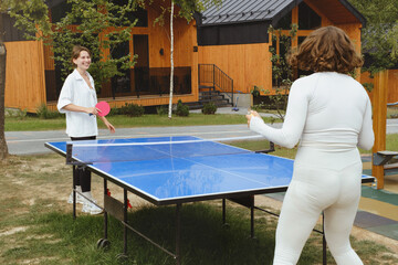 Two women play an outdoor game of table tennis, surrounded by wooden cabins and trees. Summer Sunny day. Friends having leisure activities on the fresh air.