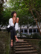 © Jesus-Salas-Dual - A young woman with curly hair sits gracefully on a brick wall in an urban park, wearing a black outfit with a white shirt draped over her shoulders. She gazes thoughtfully into the distance