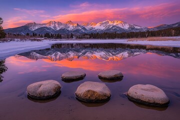 Canvas Print - Serene sunset over snow-capped mountains reflected in a calm lake with smooth rocks in the foreground.