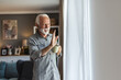 © Jelena Stanojkovic - Happy mature man relaxing at home having morning coffee looking out the kitchen window waving hello to neighbors and friends. Copy space. Lifestyle and habits.