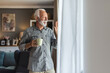 © Jelena Stanojkovic - Happy mature man relaxing at home having morning coffee looking out the kitchen window waving hello to neighbors and friends. Copy space. Lifestyle and habits.