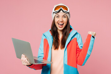 Poster - Skier young IT woman wear blue jacket ski goggles mask spend weekend winter season in mountains hold use work on laptop pc computer do winner gesture isolated on plain pink background. Hobby concept.