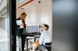 © Maskot - Businesswoman talking with male colleague sitting in wheelchair at office