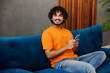 © ViDi Studio - Young smiling Indian man wear orange casual clothes sits on blue sofa couch hold use mobile cell phone stay at home hotel flat rest relax spend free spare time in living room indoor. Lounge concept.