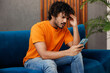 © ViDi Studio - Young sad Indian man wear orange casual clothes sit on blue sofa couch hold in hand use mobile cell phone stay at home hotel flat rest relax spend free spare time in living room indoor Lounge concept