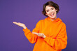 © ViDi Studio - Young smiling happy cool Caucasian woman she wear orange hoody casual clothes point hands arms aside on blank empty area mockup isolated on plain purple background studio portrait. Lifestyle concept.