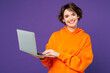 © ViDi Studio - Young smiling happy smart Caucasian IT woman wear orange hoody casual clothes hold use work on laptop pc computer looking camera isolated on plain purple background studio portrait. Lifestyle concept.