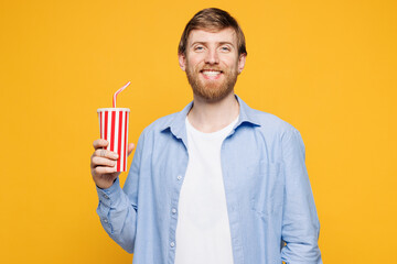 Young smiling happy man wear blue shirt t-shirt casual clothes hold in hand cup of soda pop cola fizzy water look camera isolated on plain yellow orange background studio portrait. Lifestyle concept.