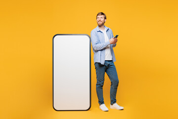 Wall Mural - Full body young smiling man wear blue shirt casual clothes stand near big huge blank screen mobile cell phone with area using smartphone isolated on plain yellow orange background. Lifestyle concept.