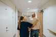 © Maskot - Rear view of female nurse with hand on shoulder of elderly man walking in illuminated corridor at care home