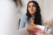 © Studio Marmellata - A young woman with curly hair wearing a light blue sweater is engaged in discussion, holding a yellow pencil in her hand while speaking in a bright and modern workspace.