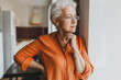 © shurkin_son - Side view of pretty charming grandmother in orange shirt and glasses leaning against wall with crossed hands next to window admiring her grandchildren playing outside, looking through with smile