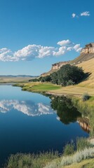 Wall Mural - Tranquil riverbank view showcases clear water reflecting hills and clouds, surrounded by lush greenery and golden grass under bright blue sky