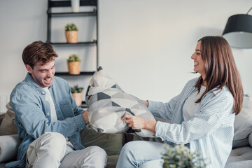 Wall Mural - Cheerful diverse married couple in love sitting on sofa in living room at home. Millennial pretty mixed race wife and Caucasian husband have fun playing fighting with pillows laughing enjoying free.