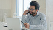 © Yuliia - Busy multitasking Arabian businessman manager consultant Indian muslim man talk mobile phone check information on laptop computer work consulting client smartphone call business consultation in office