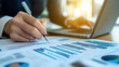 © หนึ่งฤทัย กะธง - A financial analyst reviewing business charts and graphs on paper, with an open laptop in the background, symbolizing professional work. The image focuses on the hands doing detailed ink work