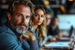 © emzee - Portrait of a man and woman in a business setting. The man has a gray beard and is looking directly at the camera. The woman is looking off to the side.  They are both seated at a table with laptops