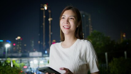 Wall Mural - Smiling woman using smartphone outdoors at night in city, surrounded by vibrant urban lights, showcasing happiness and technology in lively atmosphere. Asian Digital technology lifestyle