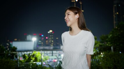 Wall Mural - Confident woman enjoys serene night outdoors, wearing wireless earbuds while taking in vibrant urban skyline of city. Asian Digital technology lifestyle